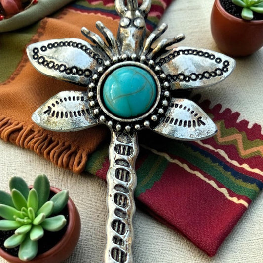 Decorative silver and turquoise dragonfly purse charm on a colorful fabric background with potted plants.