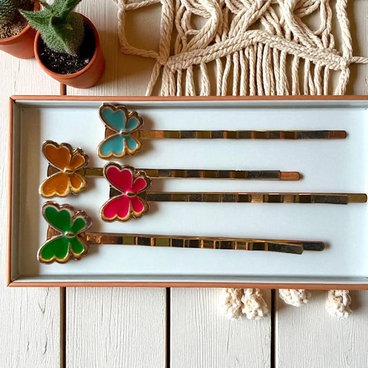 Set of colorful butterfly-shaped hair pins in a box on a wooden surface with plants and macrame wall hanging.