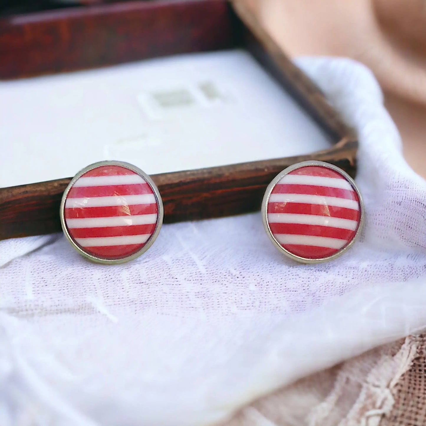 Round red and white striped stud earrings in a silver-tone setting displayed near a rustic tray