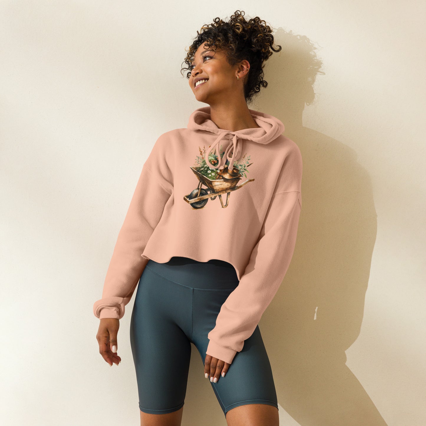 Model wearing peach cropped hoodie with gardening wheelbarrow graphic