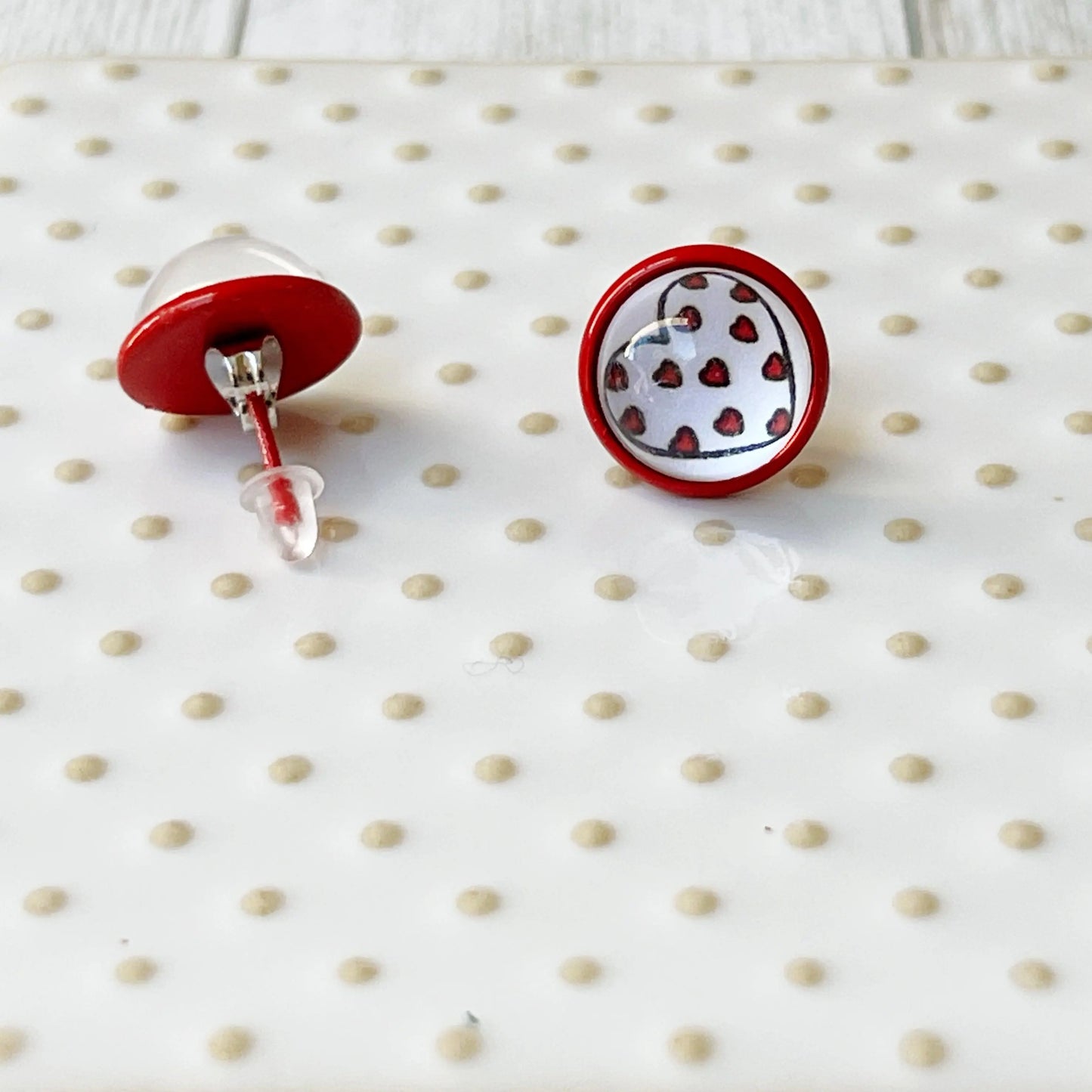 Back of round red stud earrings with white heart artwork and tiny red heart details displayed on a glass tray