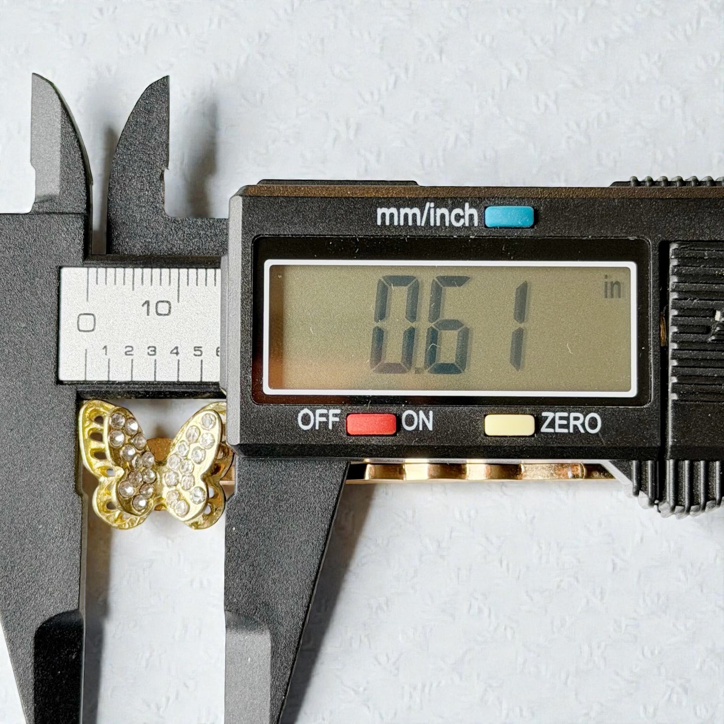 Digital caliper measuring a gold butterfly pendant with digital display showing measurement.