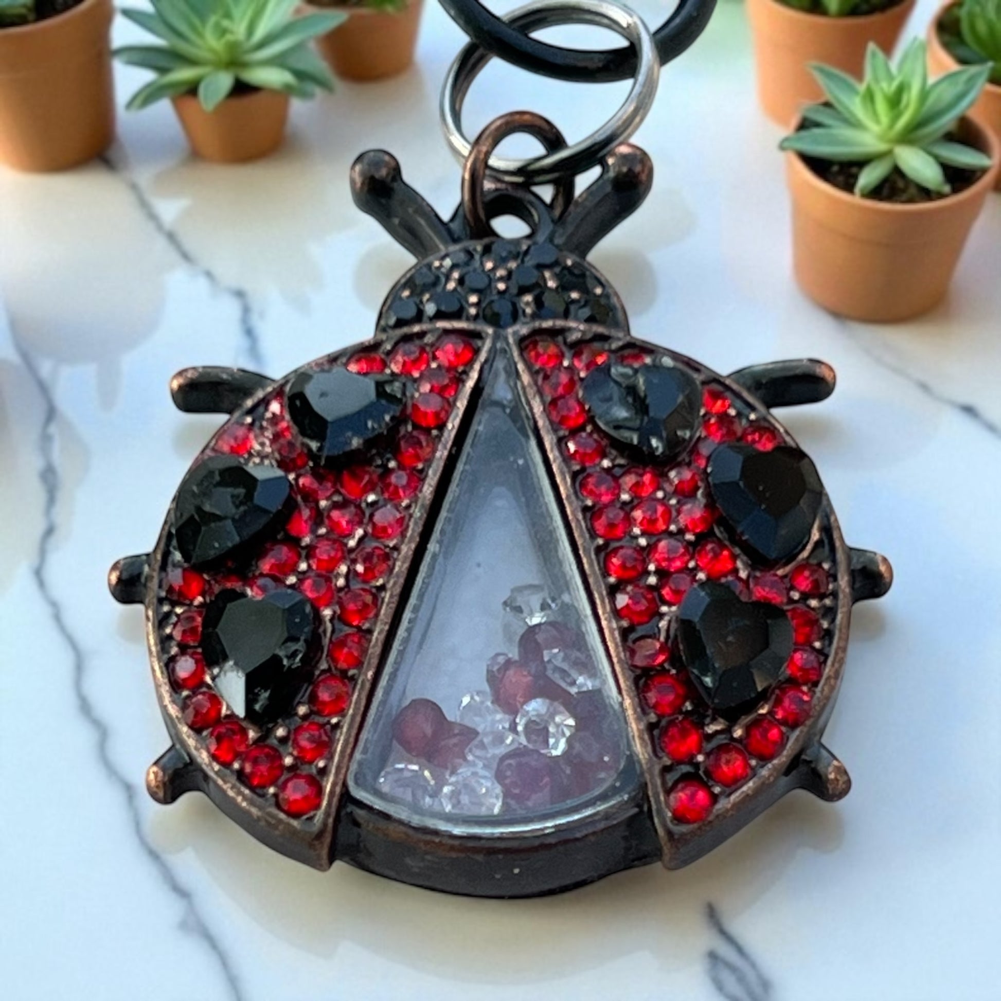 Decorative ladybug keychain with red and black gemstones on a marble surface with potted plants in the background.