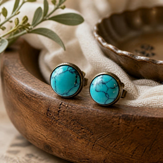 One pair of antique brass-tone 12 mm faux-turquoise round stud earrings resting on cream linen by a rustic jewelry dish