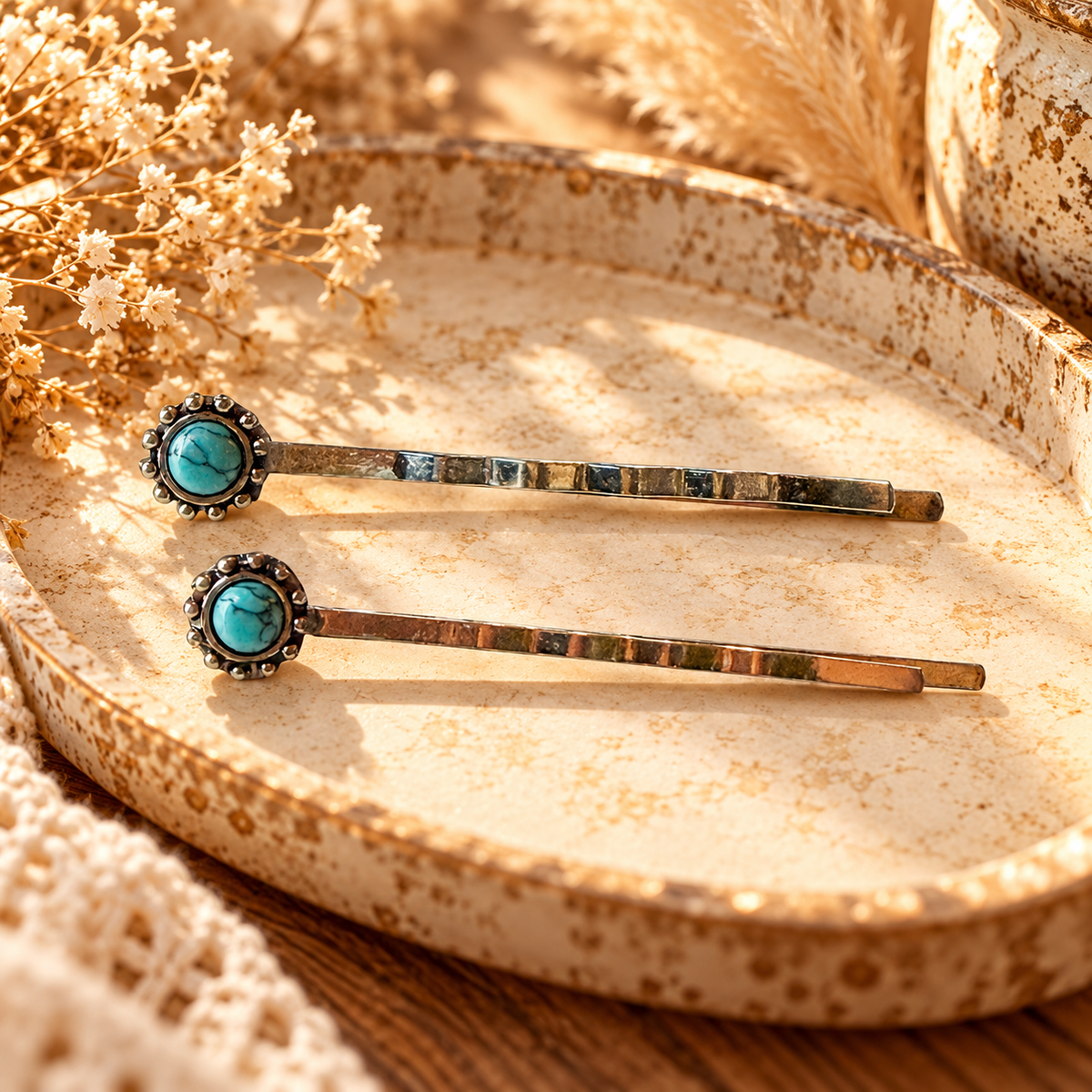 Exactly two silver-tone faux-turquoise western hair pins on a warm ivory stone tray with dried flowers; styled illustration