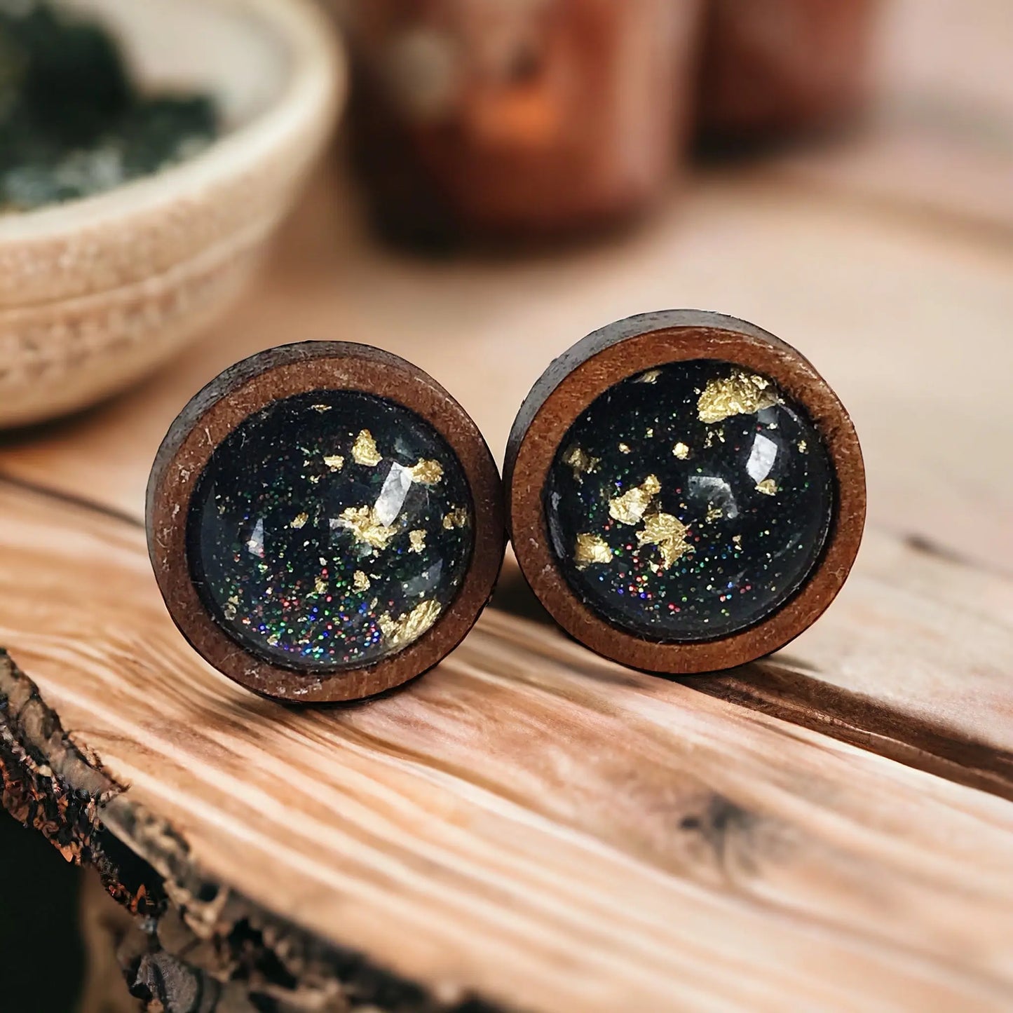 Round black resin wood stud earrings with gold flake detail and subtle sparkle on a whitewashed wood background