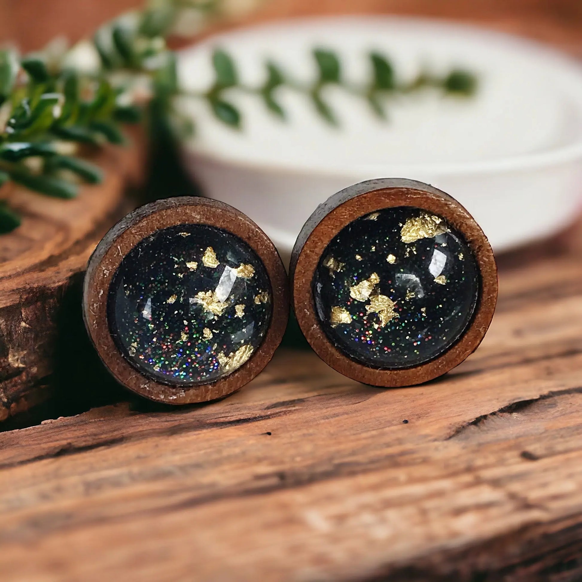 Round black resin wood stud earrings with gold flake detail and subtle sparkle on a decorative wood background
