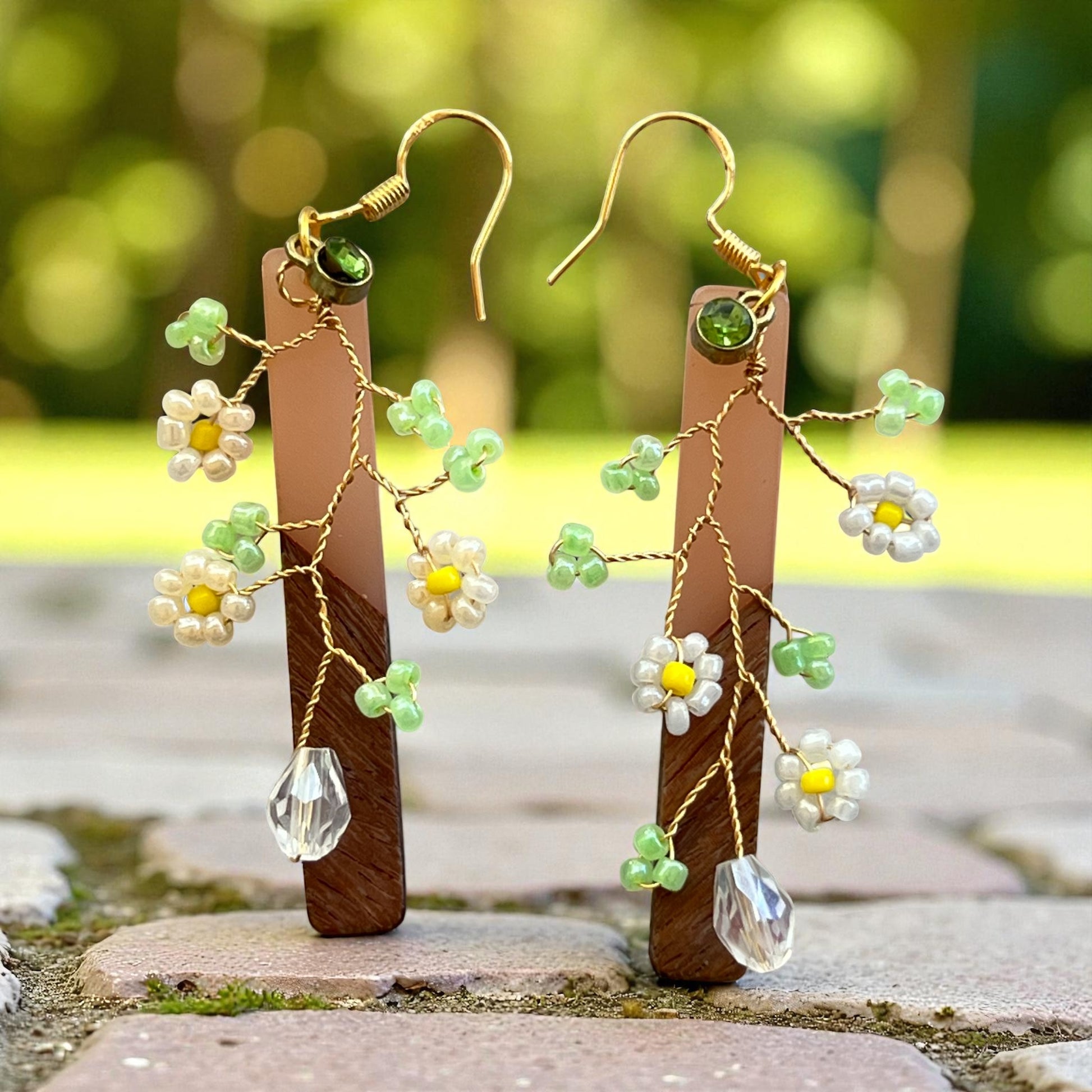 Pair of floral earrings with green beads and clear crystals with wood bars against a blurred natural background.