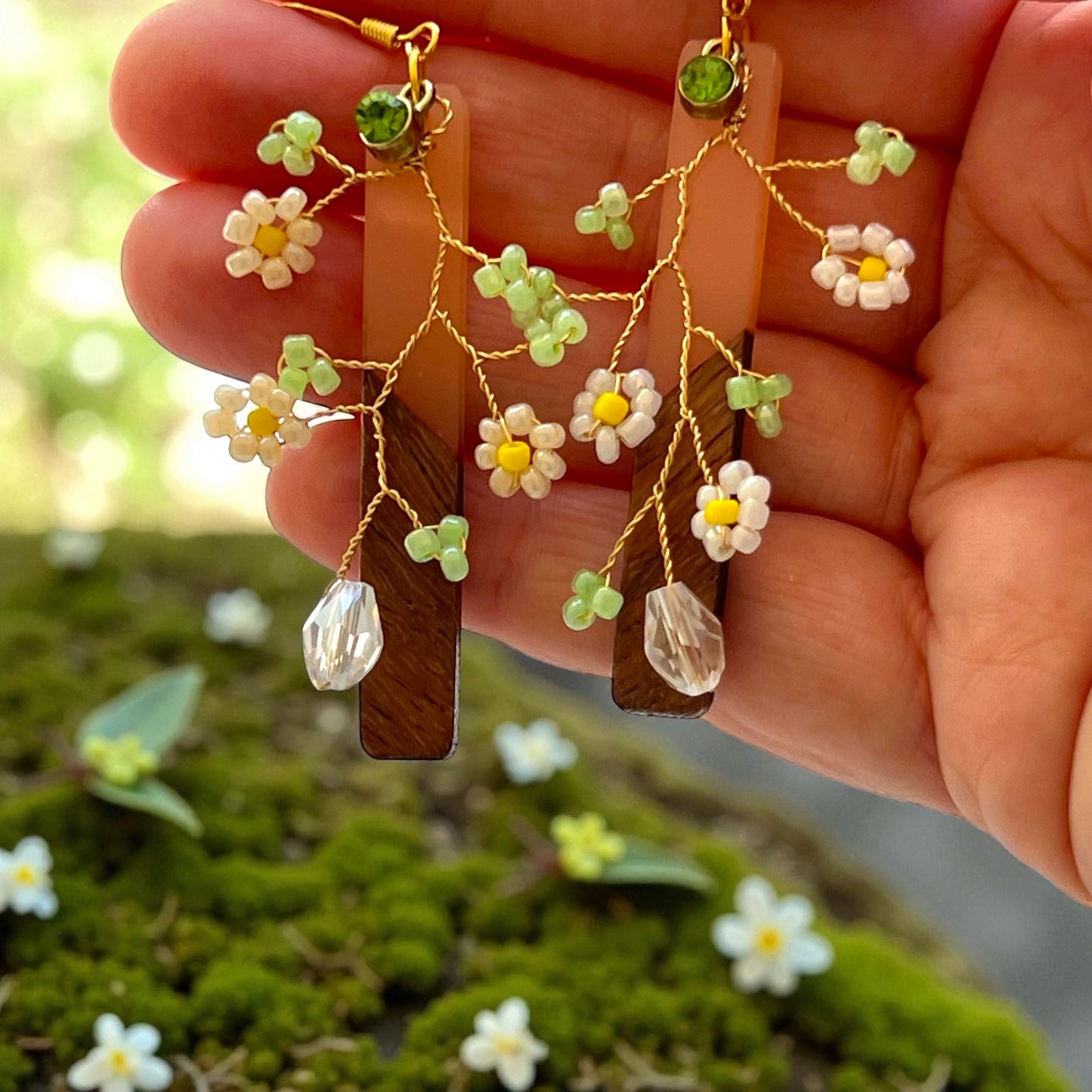 Hand holding a pair of floral earrings with green leaves and wooden accents against a blurred natural background.