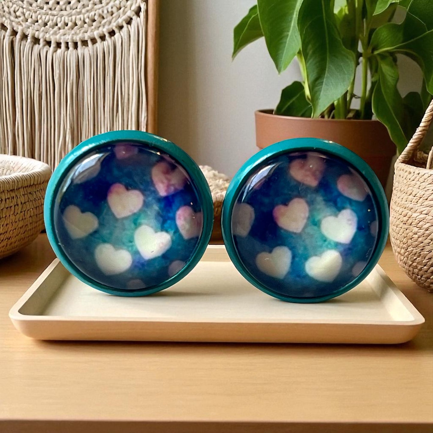 Two blue earrings with heart patterns on a white tray, surrounded by decorative items.