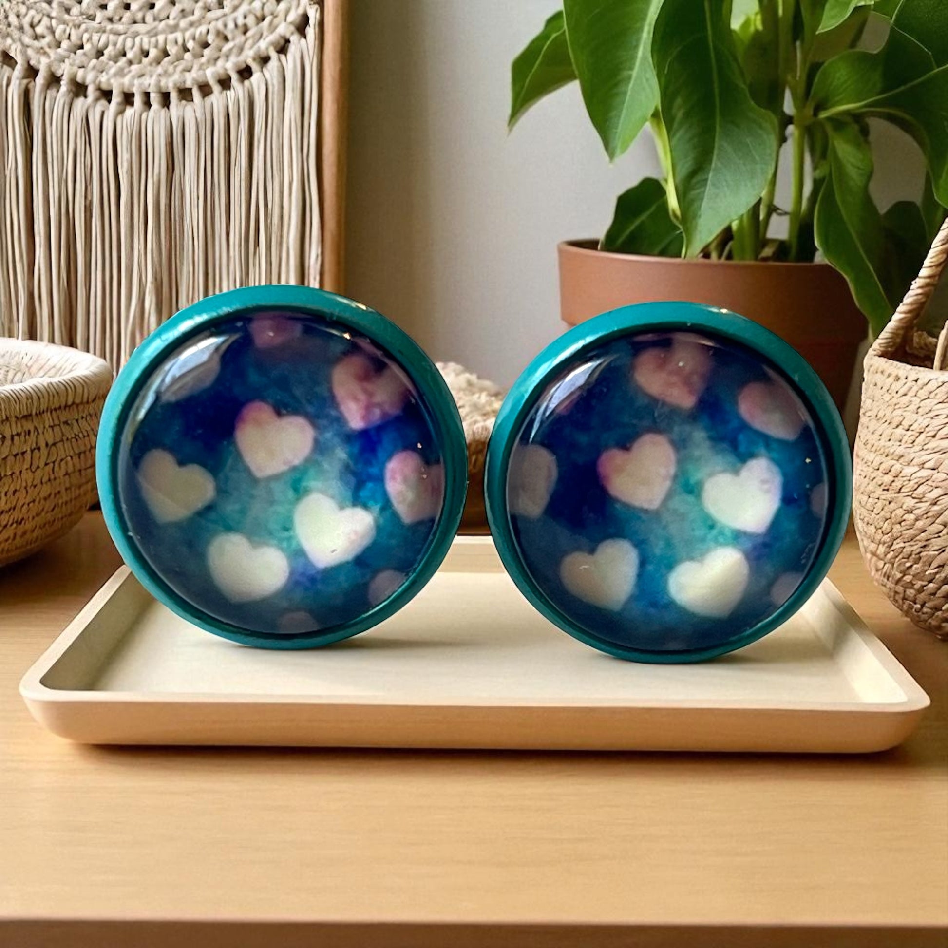 Two blue earrings with heart patterns on a white tray, surrounded by decorative items.