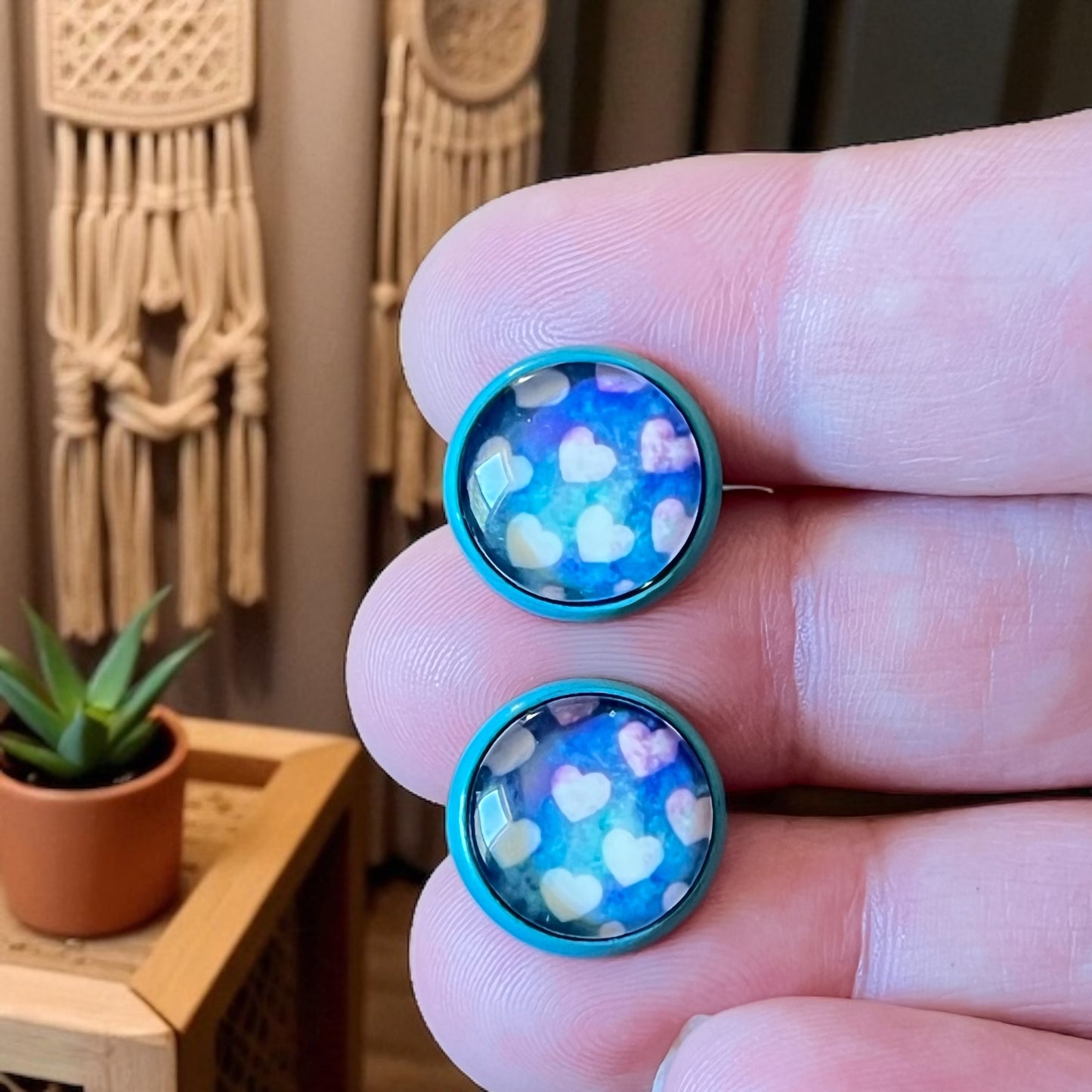 Close-up of a hand holding two blue earrings with heart pattern, blurred indoor background