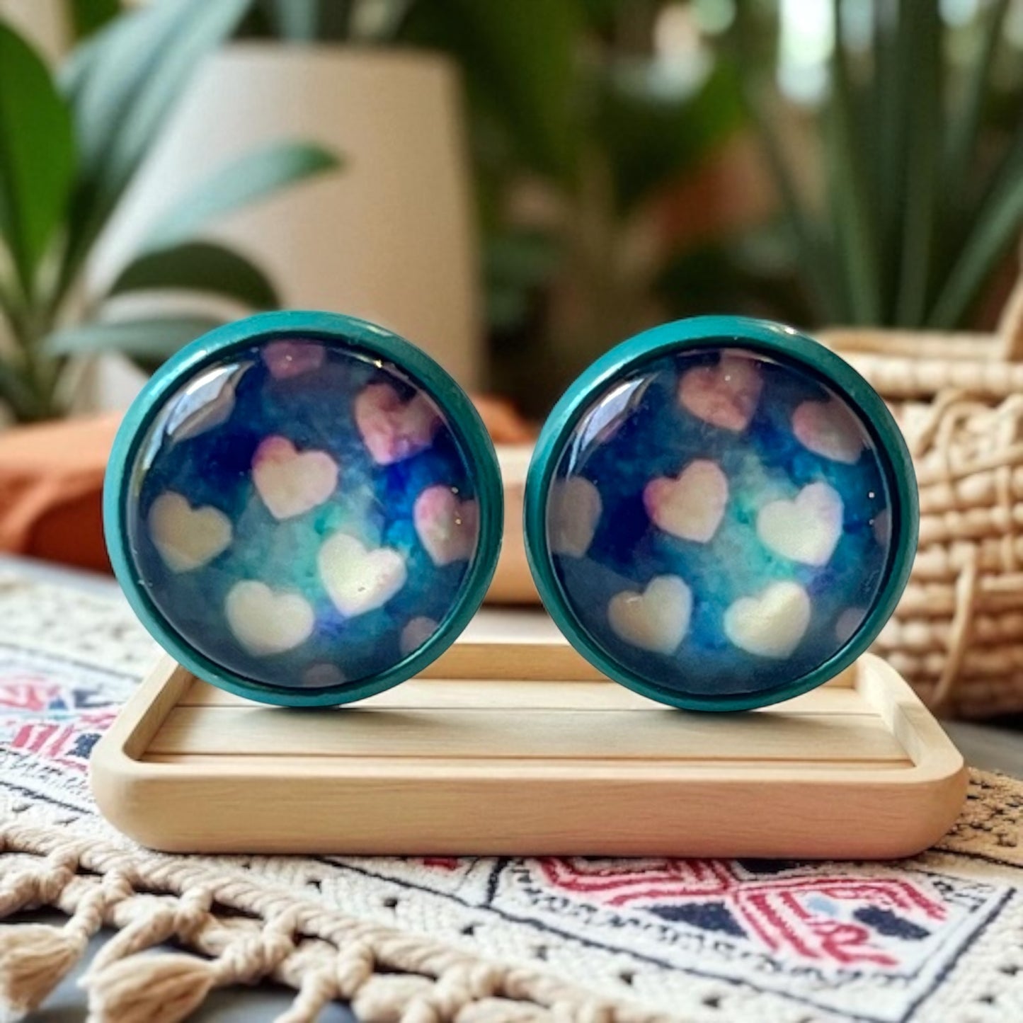Pair of blue earrings with heart pattern on a wooden stand with plants in the background