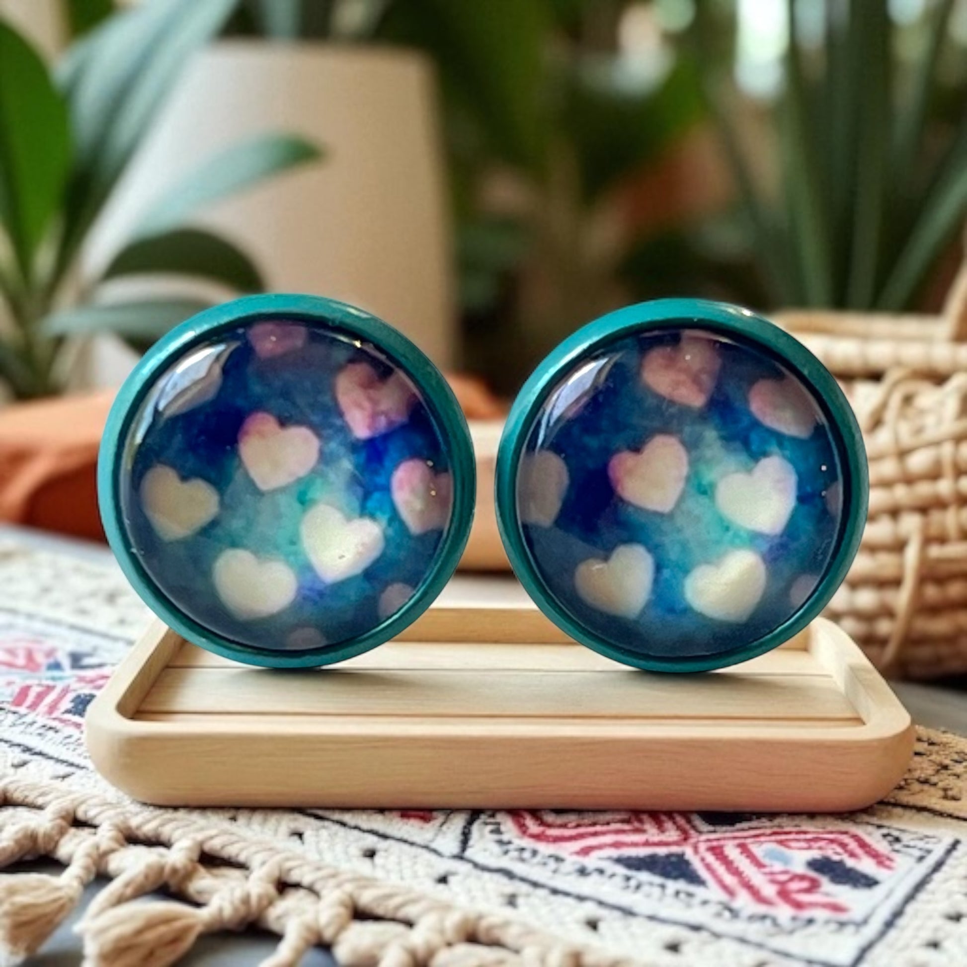 Pair of blue earrings with heart pattern on a wooden stand with plants in the background