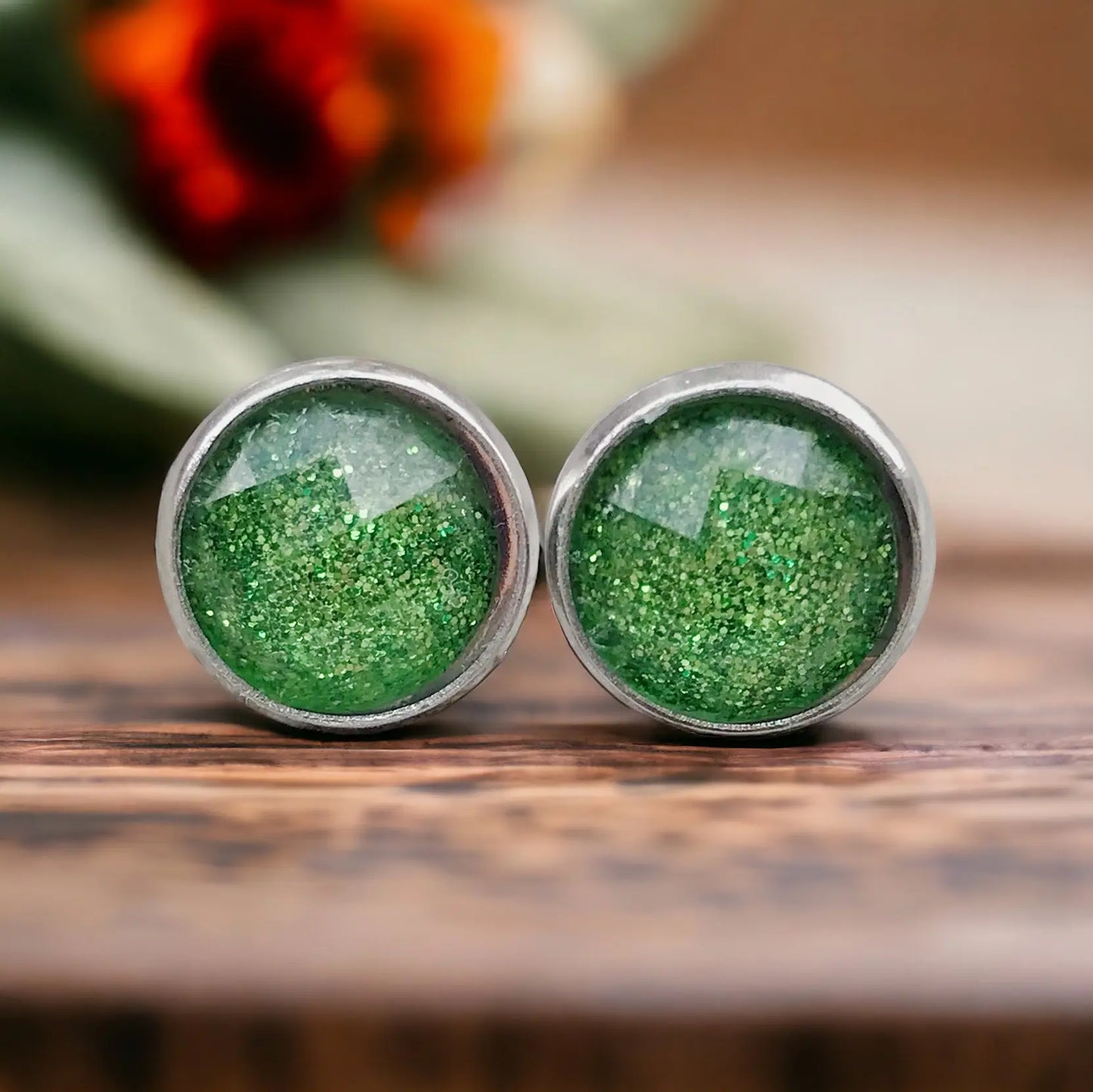 Green glittery stud earrings on a wooden surface with a blurred floral background