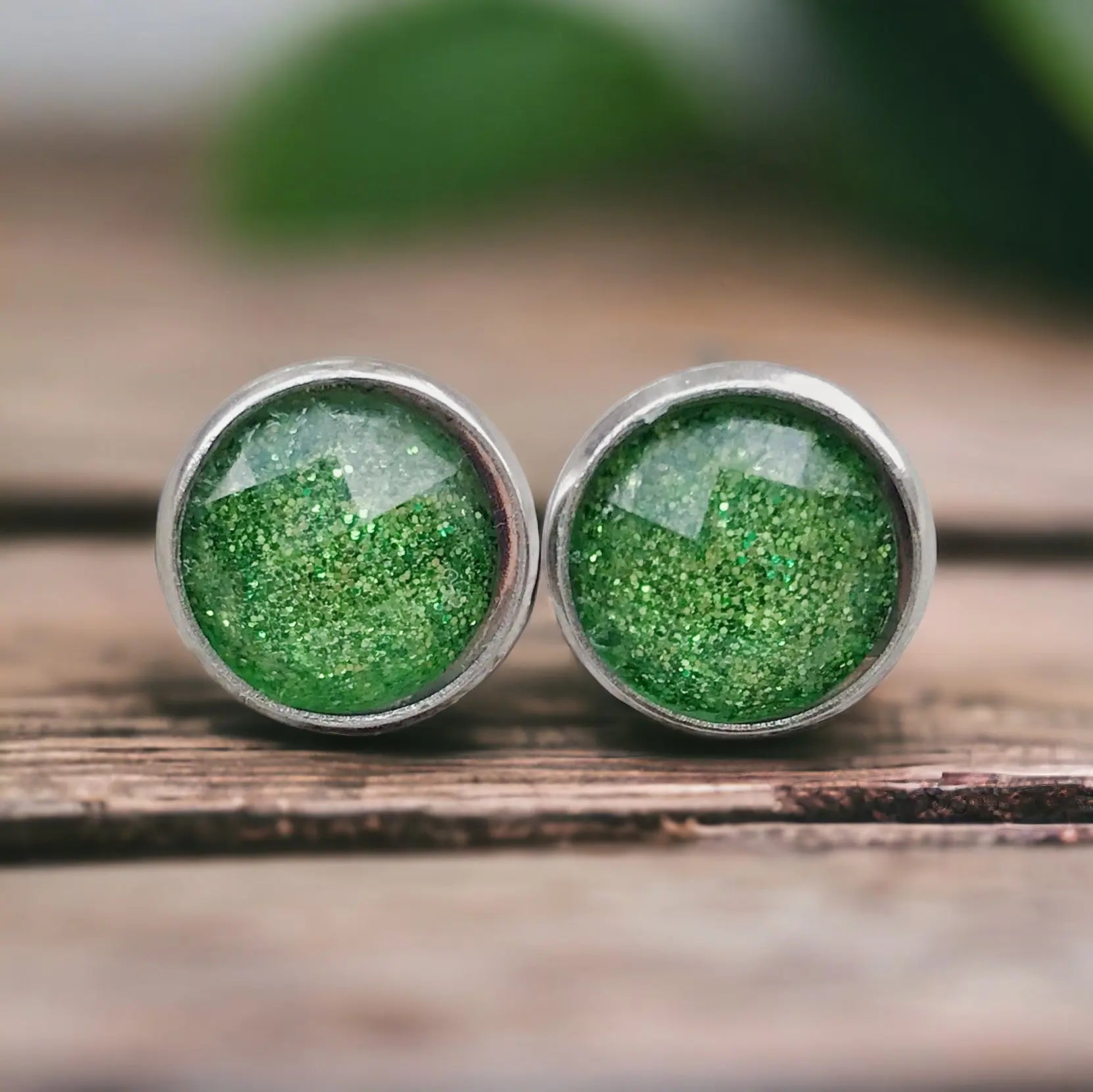 Green glittery stud earrings on a wooden surface with a blurred natural background