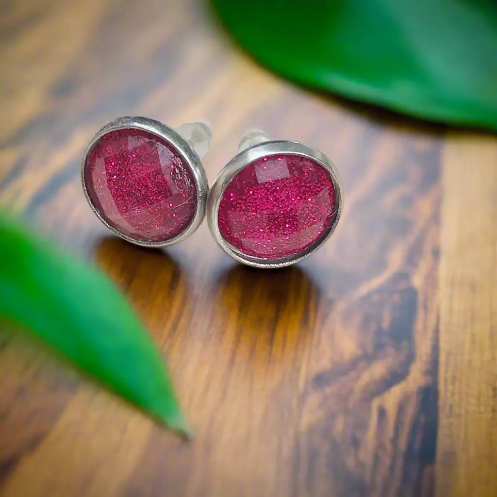 Pink glittery stud earrings on a wooden surface with green leaves.