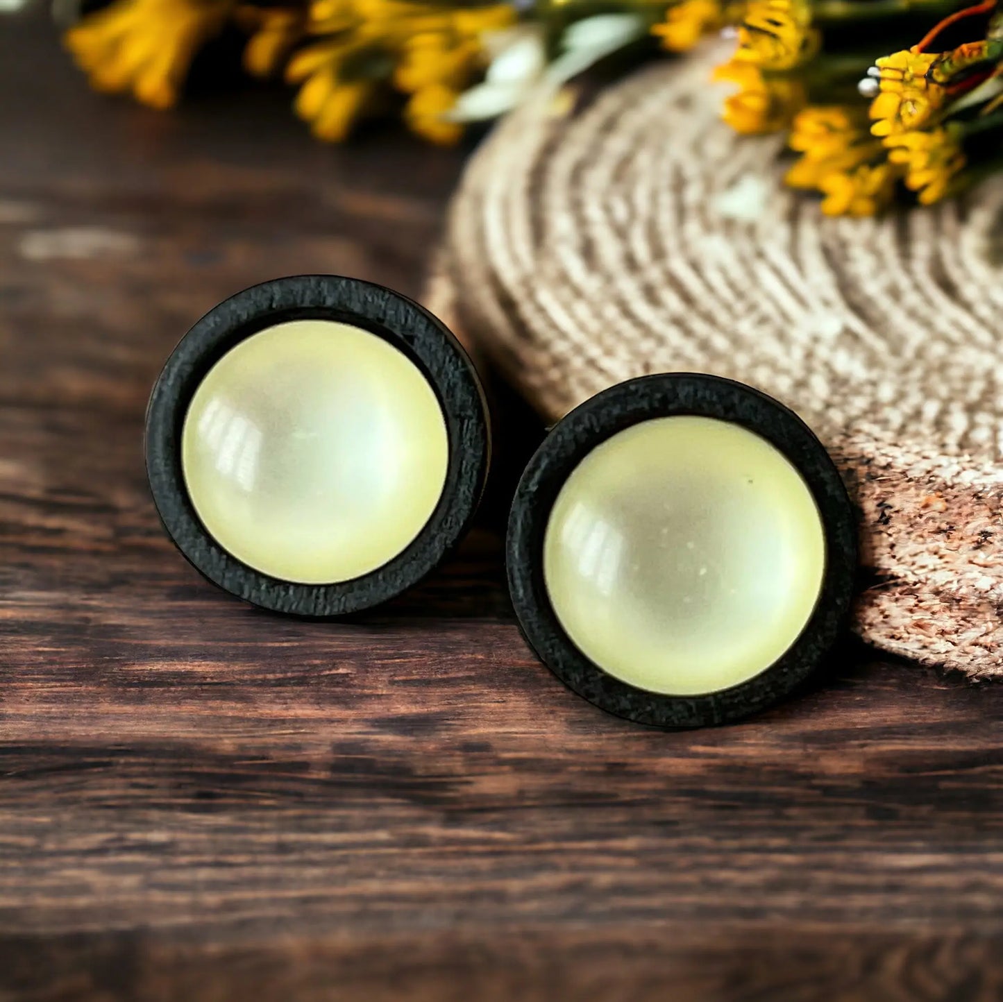 Pair of round yellow earrings with black rims on a wooden surface with flowers in the background