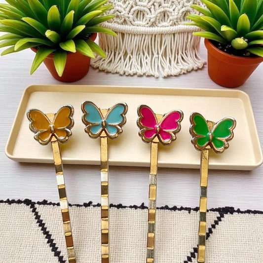 Four butterfly-shaped hairpins with gold bases on a beige tray, surrounded by potted plants.