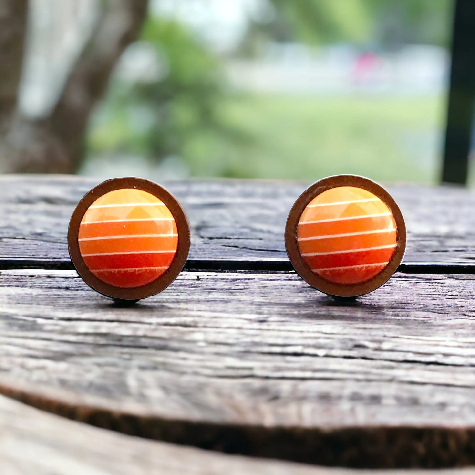 Round orange gradient striped wood stud earrings displayed on a rustic dark wood surface