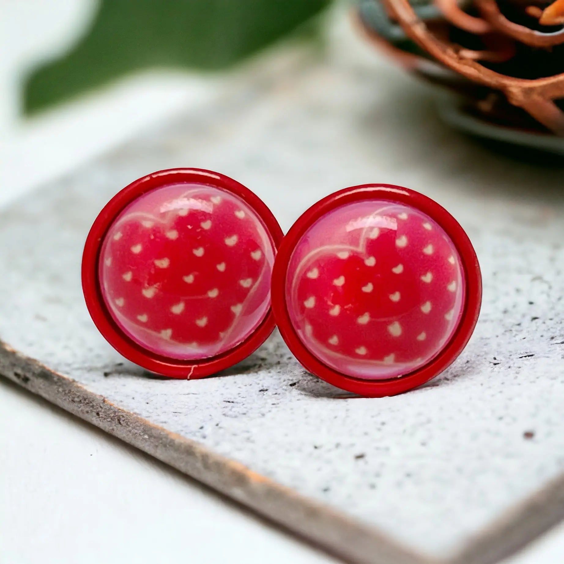 Round red stud earrings with tiny white heart pattern in a red setting displayed on a light stone surface