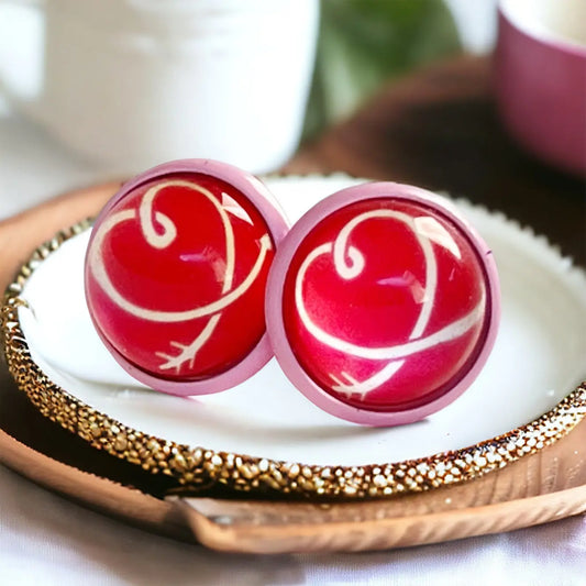 Round pink stud earrings with red and white heart artwork displayed on a decorative tray