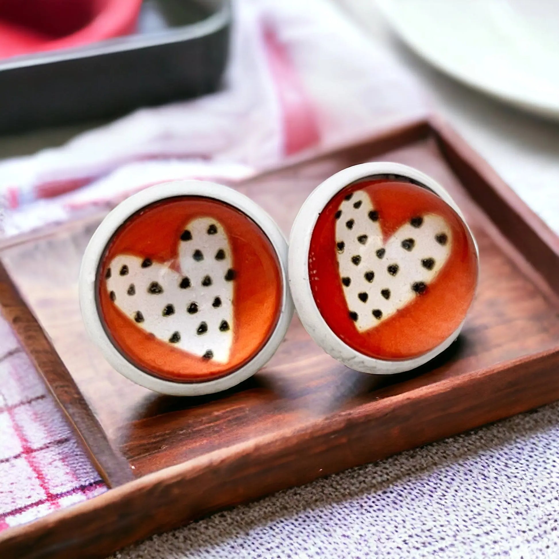 Round red stud earrings with white polka dot heart design in a silver-tone setting displayed on a wooden tray