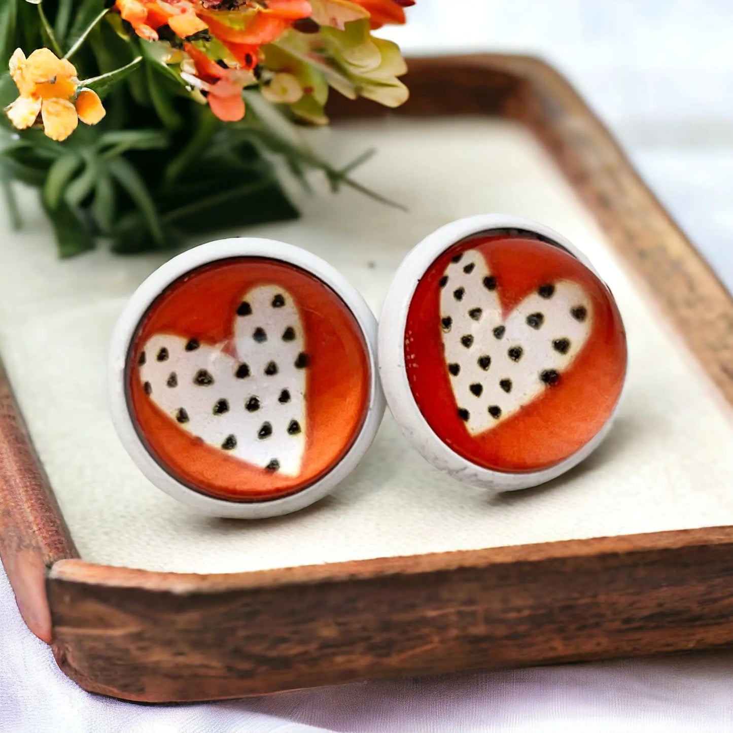 Heart-shaped earrings with white pattern on red background on a wooden tray with flowers in the background