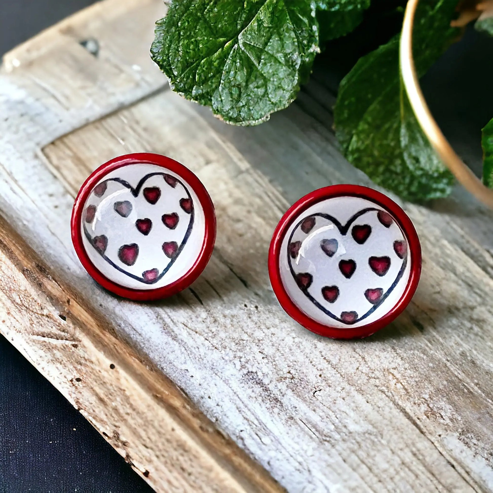 Round red stud earrings with white heart artwork and tiny red heart details displayed on a boho wooden tray