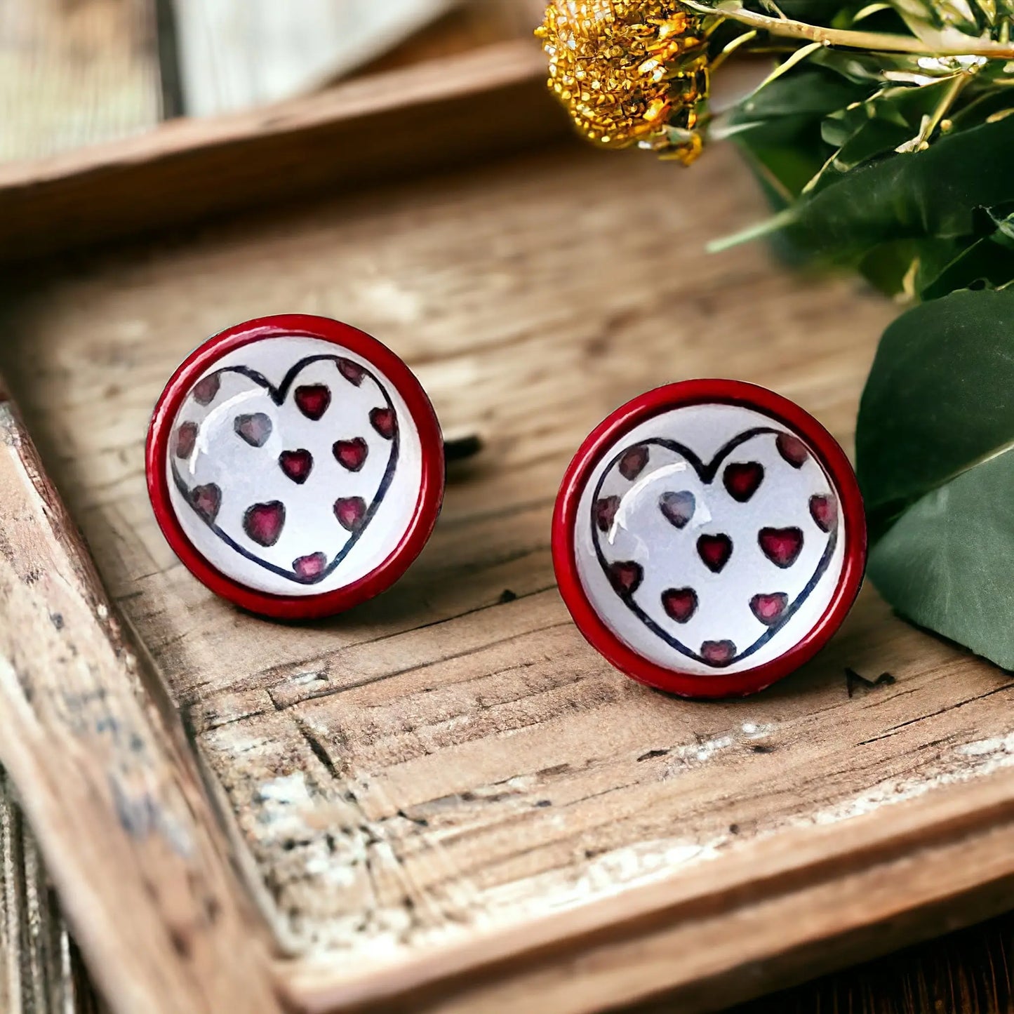 Round red stud earrings with white heart artwork and tiny red heart details displayed on a wooden tray
