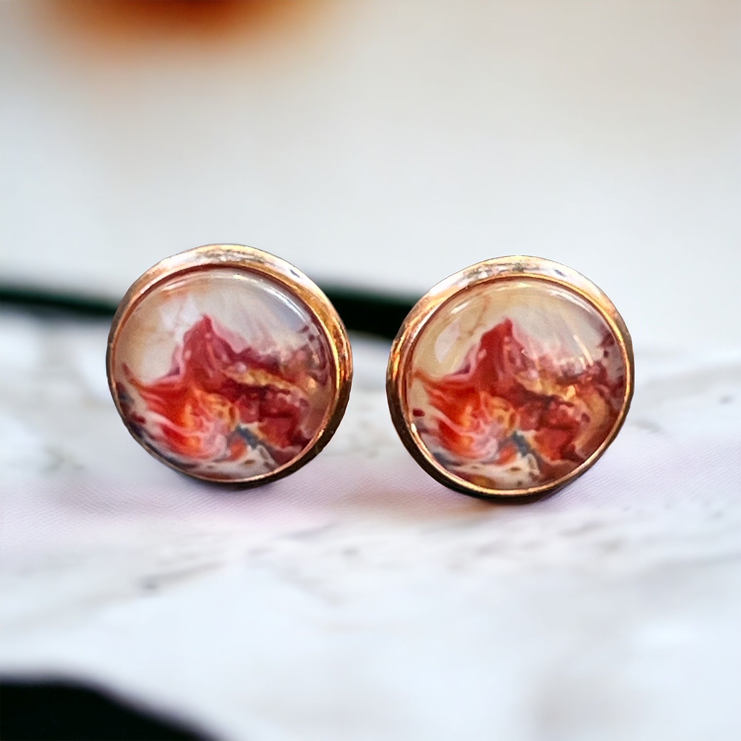 Rose gold setting stud earrings with glossy marbled resin in red, blush, and white on blurred marble background