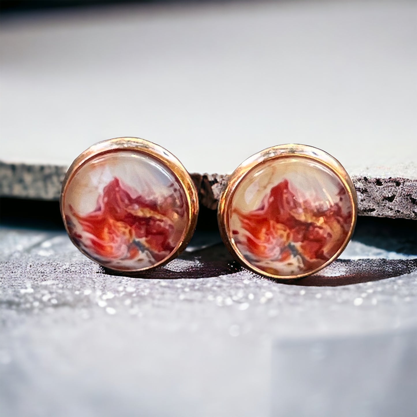 Rose gold setting stud earrings with glossy marbled resin in red, blush, and white resting on rock slat