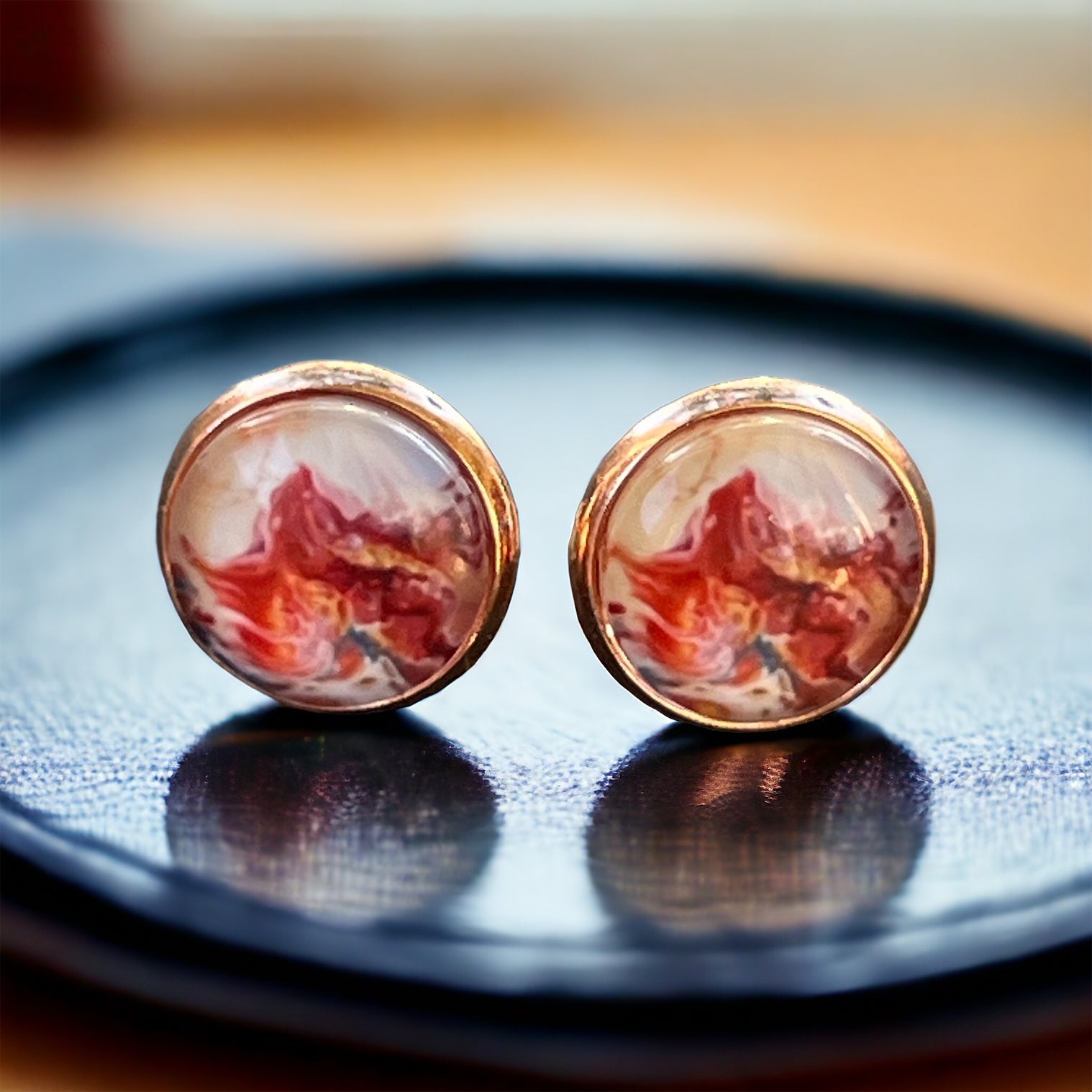 Rose gold setting stud earrings with glossy marbled resin in red, blush, and white on reflective tray