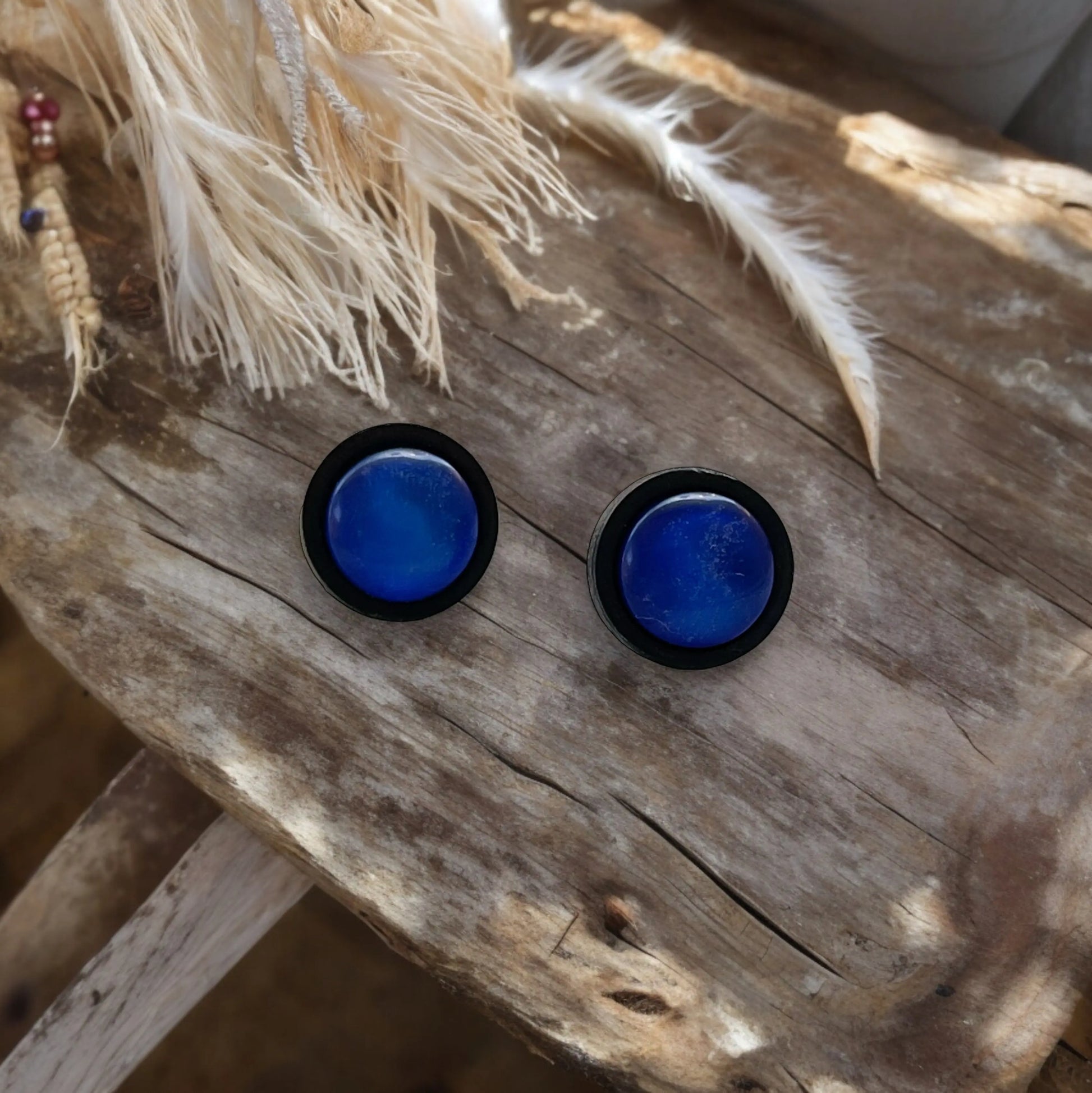 Blue circular earrings with black borders on a wooden surface with feathers.