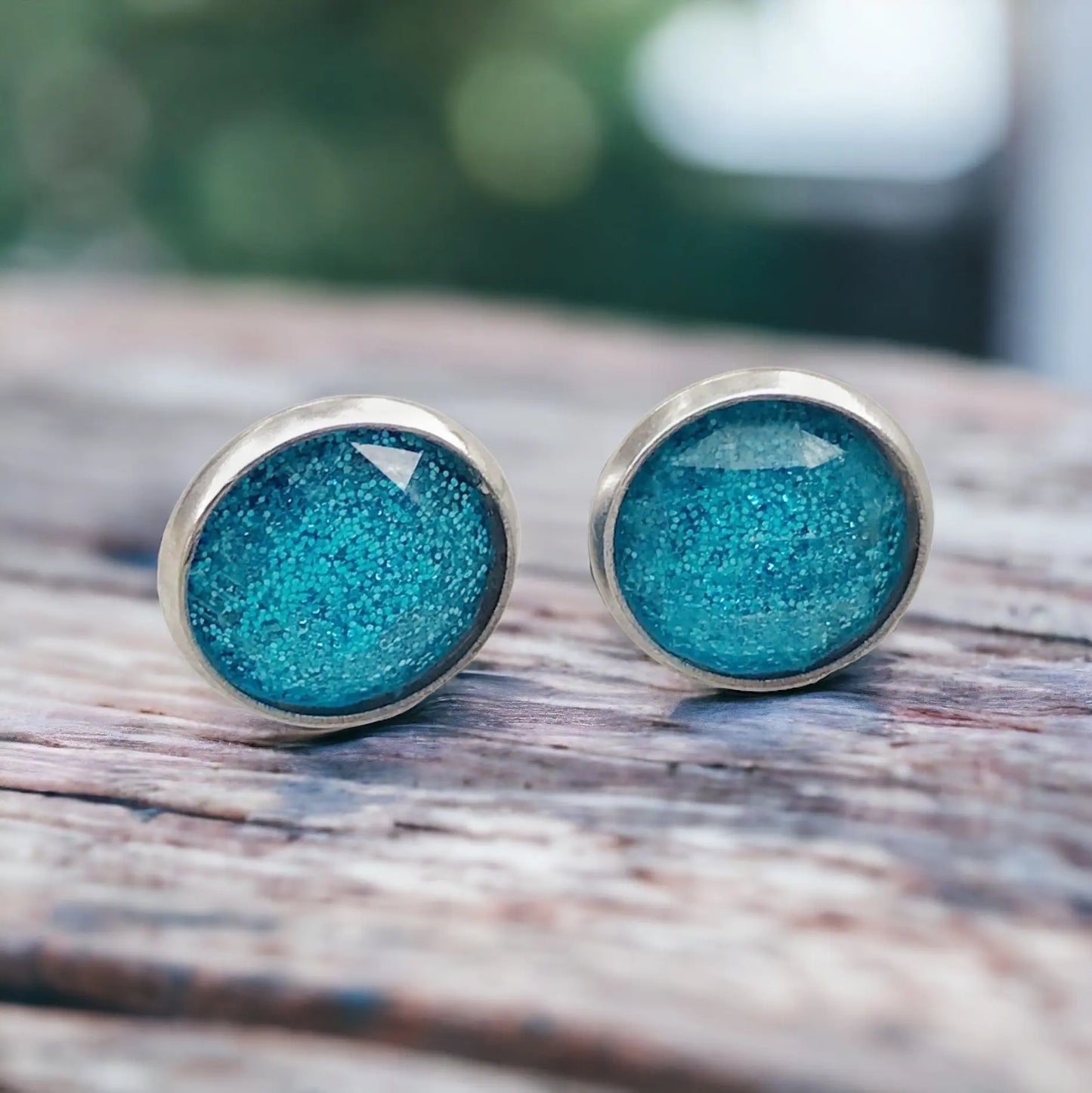 Round sky blue glitter stud earrings in a silver-tone setting displayed on a light wood surface
