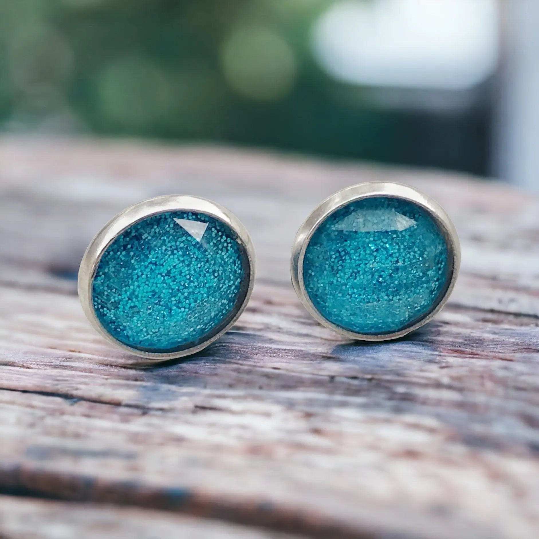 Round sky blue glitter stud earrings in a silver-tone setting displayed on a light wood surface
