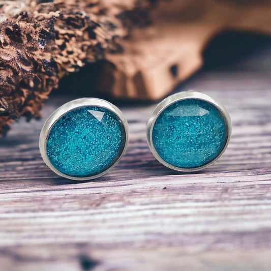 Round sky blue glitter stud earrings in a silver-tone setting displayed on a rustic wood surface