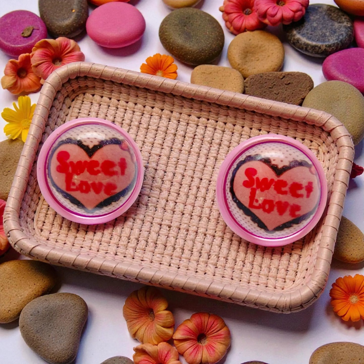 A pair of 'Sweet Love' heart-shaped earrings on a woven tray with colorful stones and flowers in the background.