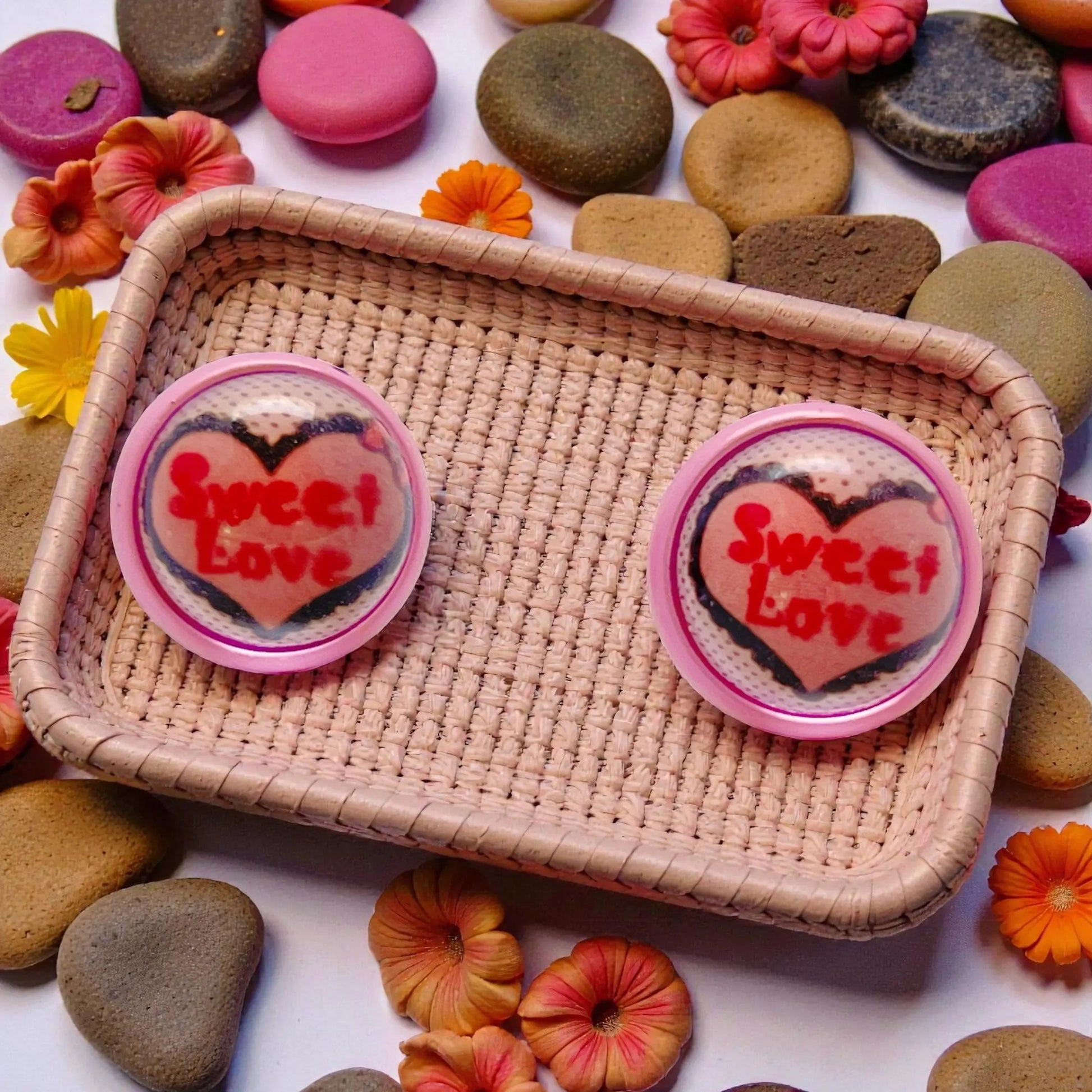 A pair of 'Sweet Love' heart-shaped earrings on a woven tray with colorful stones and flowers in the background.