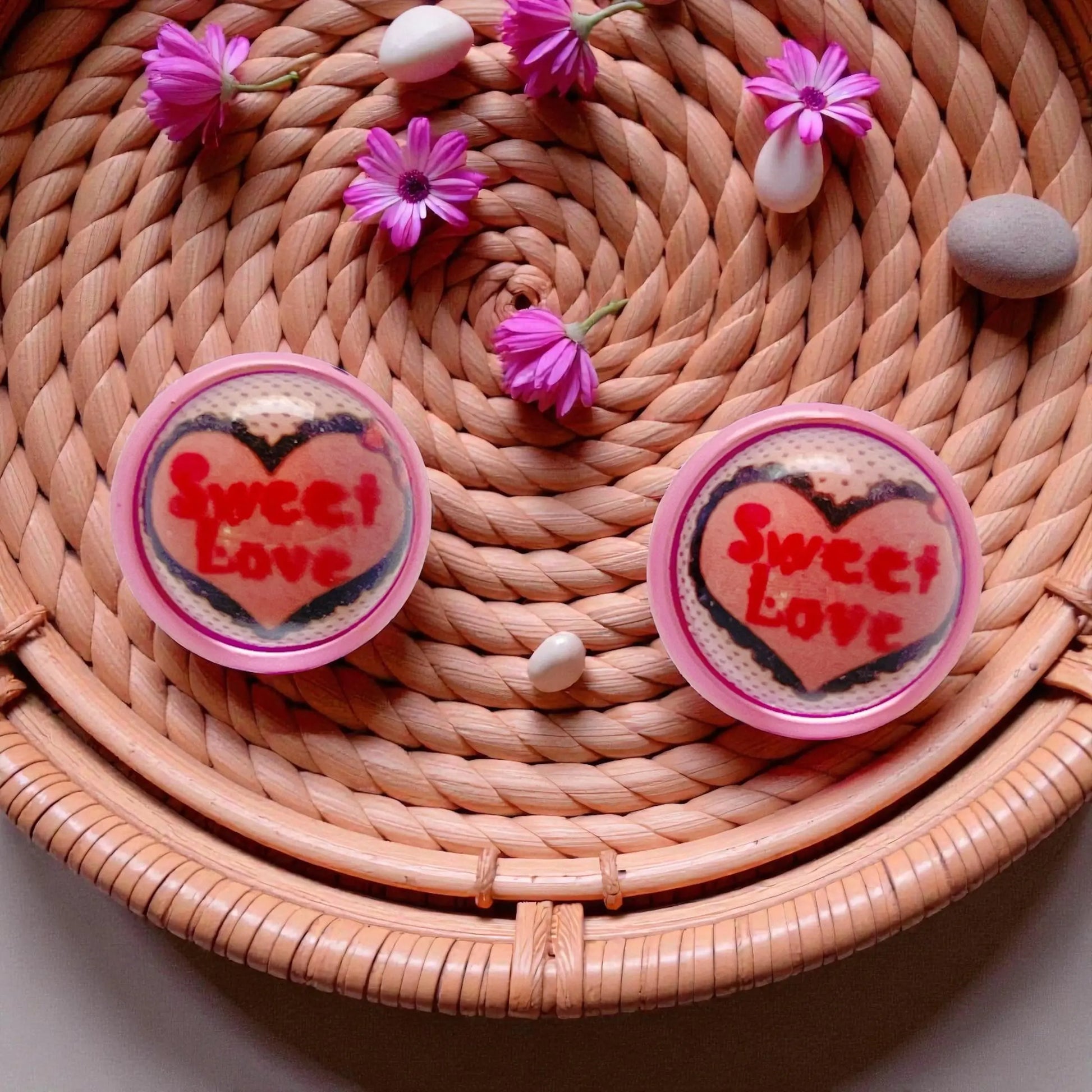 Two heart-shaped 'Sweet Love' earrings on a woven surface with pink flowers.