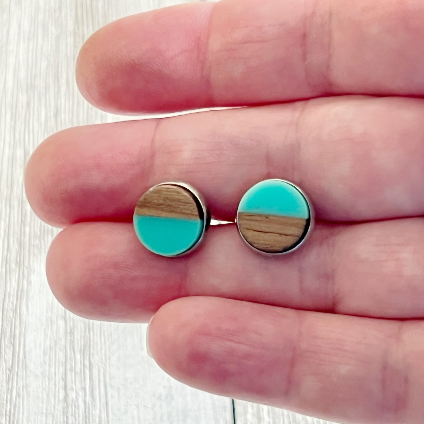 Round teal and wood stud earrings with a glossy resin top displayed in hand to show scale 