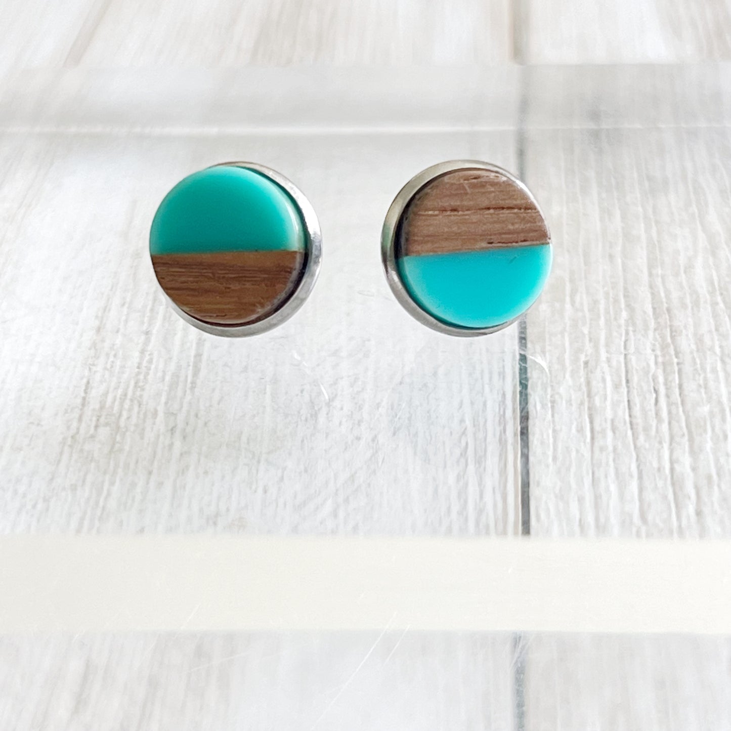 Round teal and wood stud earrings with a glossy resin top displayed on a clear riser