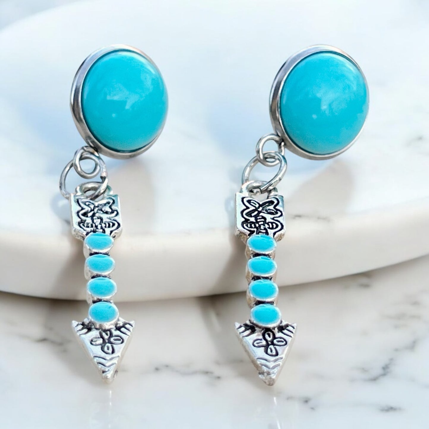 Turquoise arrow dangle earrings with faux turquoise stud tops and silver-tone arrow charms On white tray with marbled background 