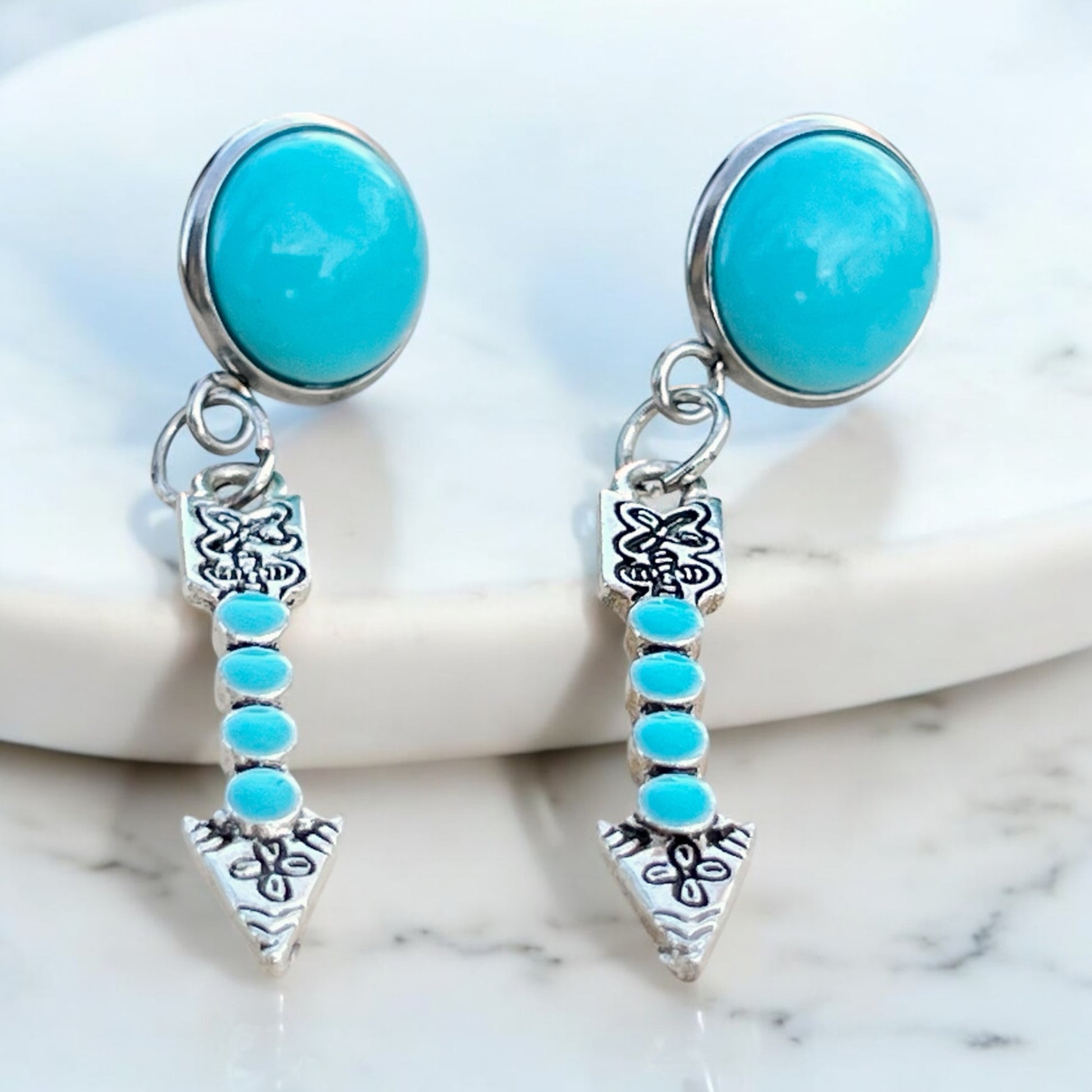 Turquoise arrow dangle earrings with faux turquoise stud tops and silver-tone arrow charms On white tray with marbled background 
