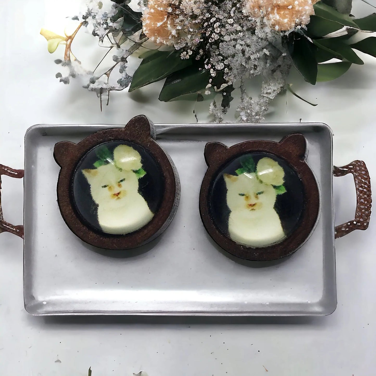 Two cat-shaped earrings on a tray with flowers in the background