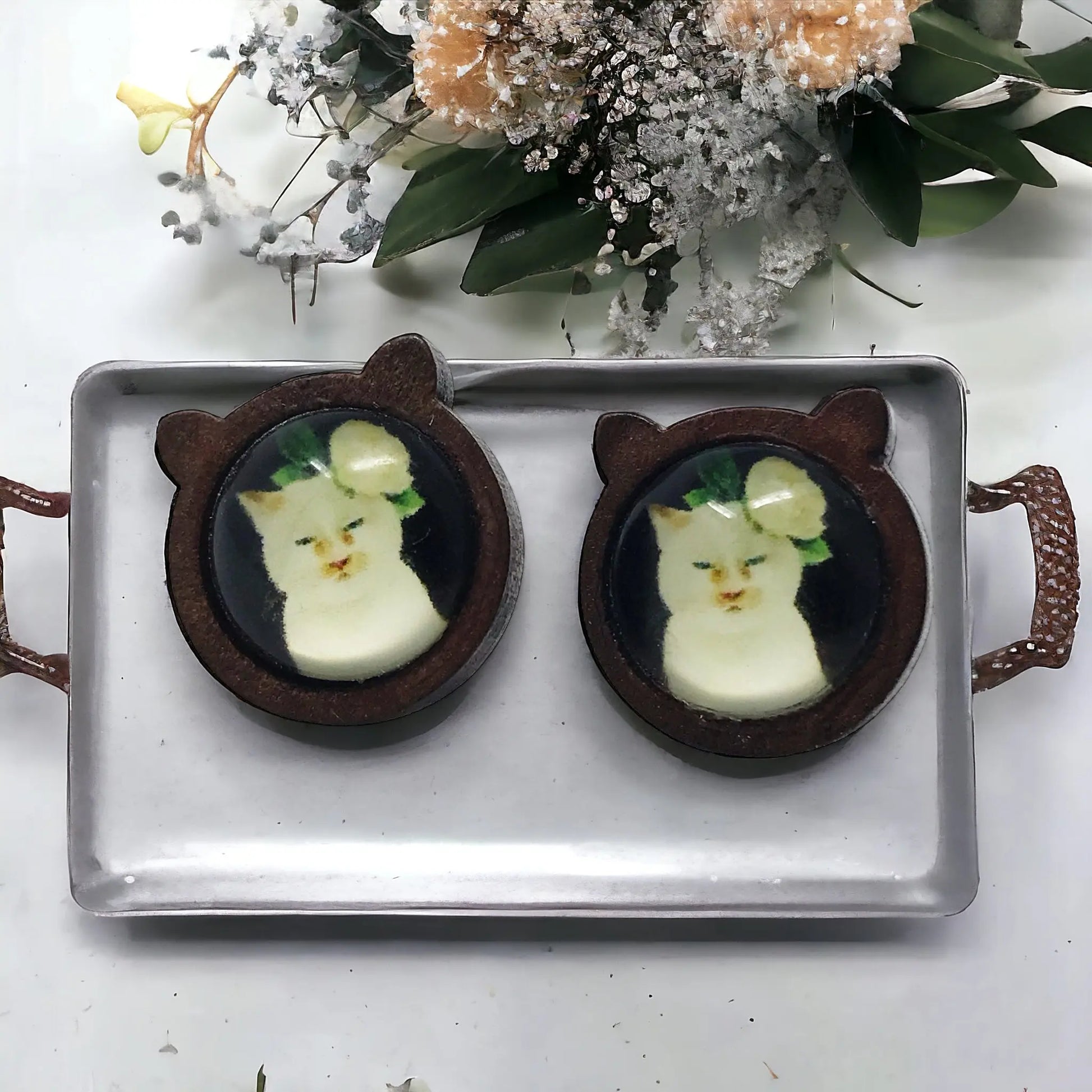Two cat-shaped earrings on a tray with flowers in the background