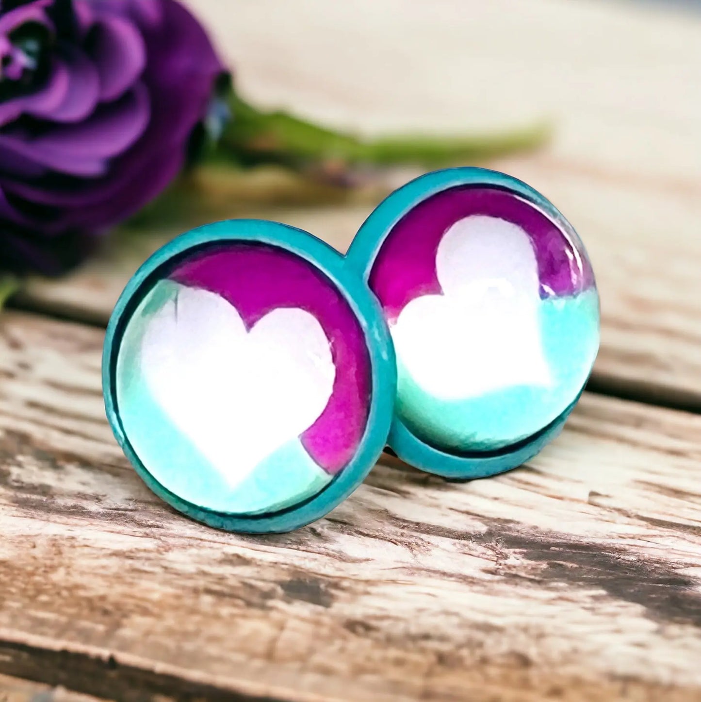 Round teal and pink stud earrings with white heart artwork displayed on a rustic wood piece