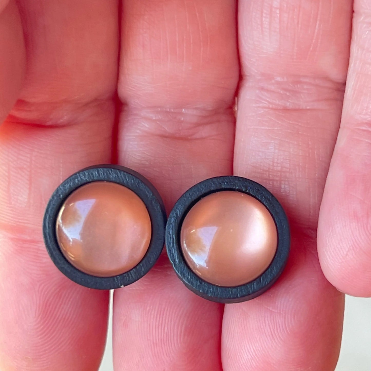 A pair of earrings with a beige center and black outer rim held between fingers.