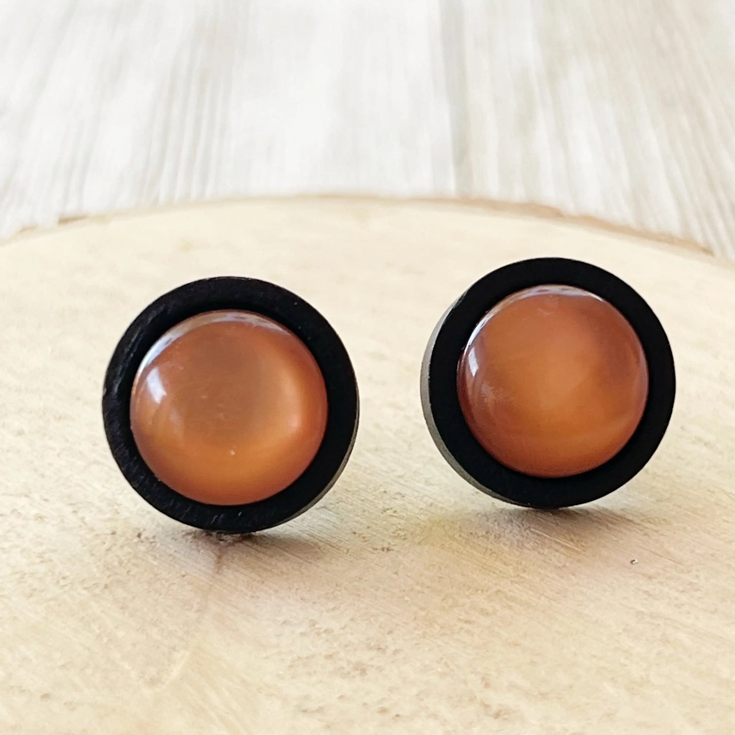 Round caramel brown resin stud earrings with black wood border displayed on a wood slice
