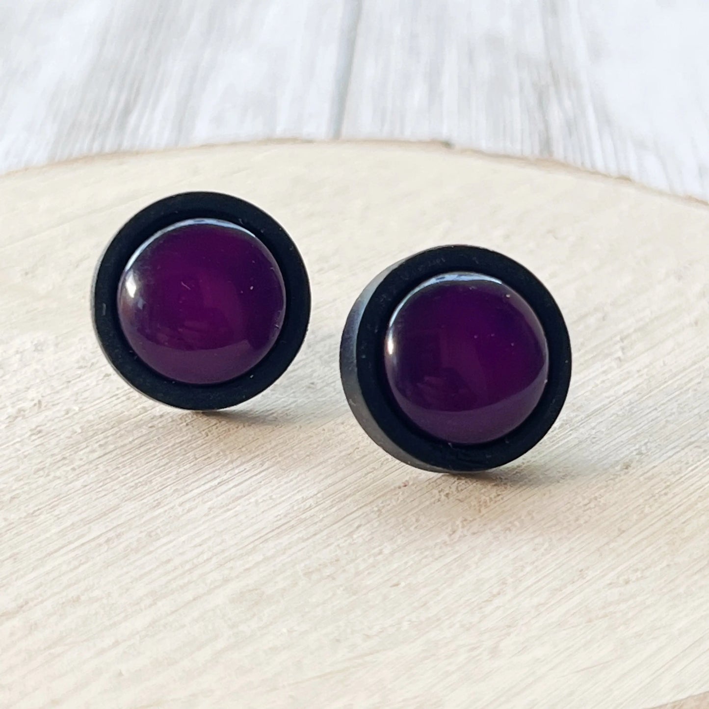Round dark purple resin stud earrings with black wood borders displayed on a light wood surface
